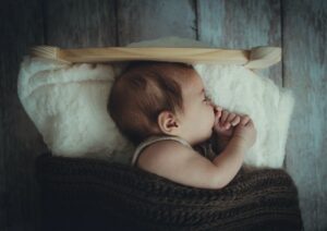 Would you consider renting the baby clothes for a newborn?-Baby sound a sleep in a wooden bed and a white pillow and brown knitted blanket.