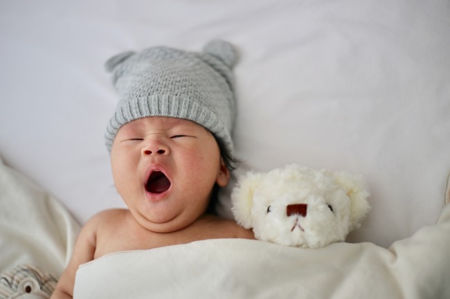 5 benefits organic baby clothes have?-Yawning baby in bed with soft toy and grey hat on.